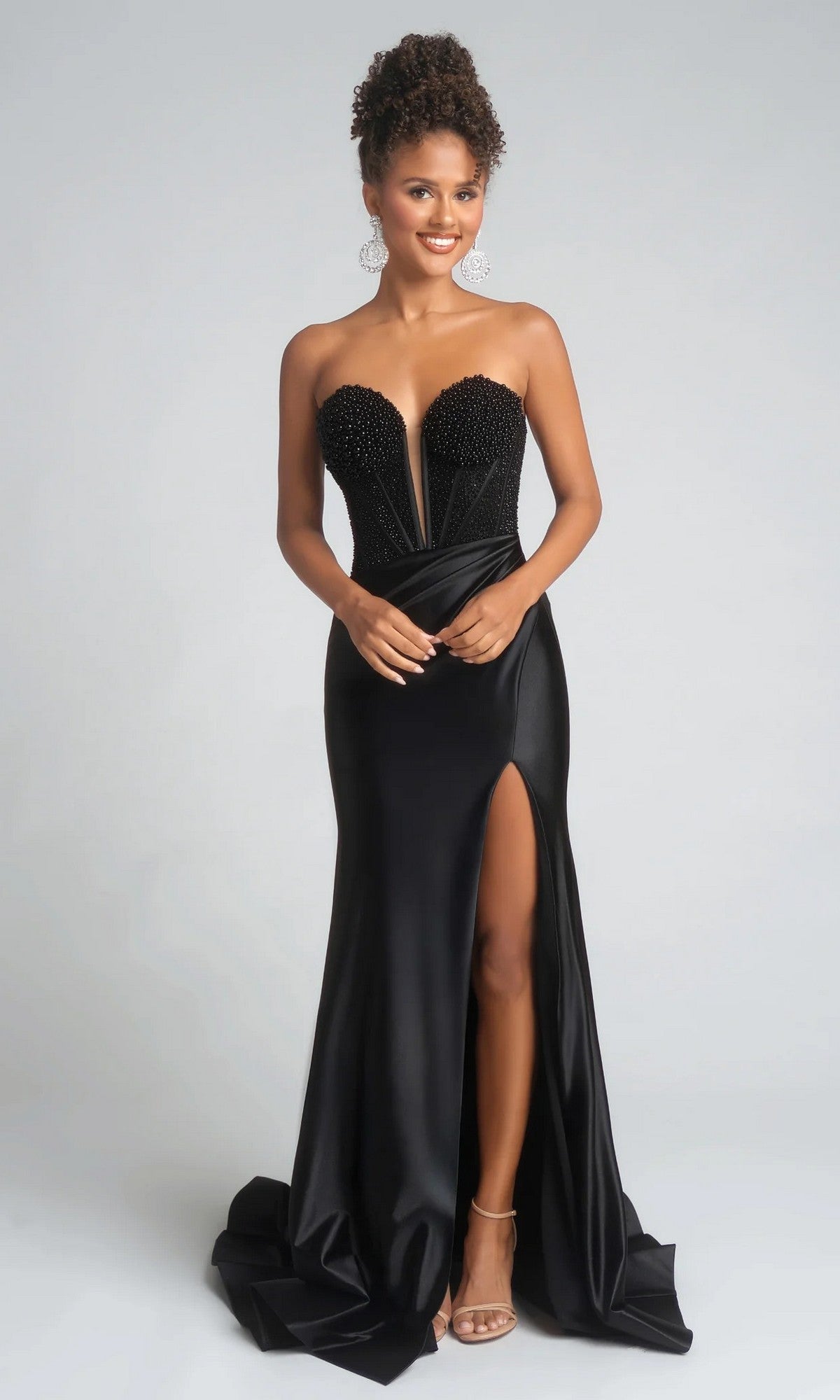 A woman in the Portia and Scarlett PS26039 long prom dress featuring a strapless beaded corset bodice, sweetheart neckline, thigh-high slit, and open-toe heels stands against a plain background.